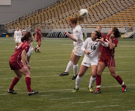 Color photograph of two soccer teams playing each other. The women wear white and red uniforms. In the background are empty stadium stands.